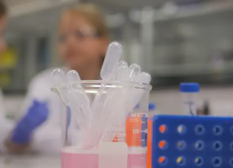 A close up of some pipettes and a test tube holder. In the background a child in a lab coat