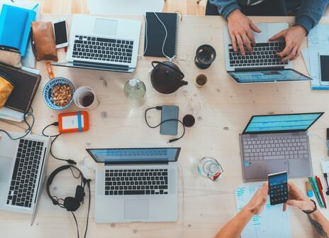 A table top is filled with laptops, chargers, phones and cups of tea and coffee.