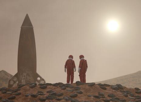 Two astronauts and a rocket are standing on the surface of a planet. It is dusty and red. 