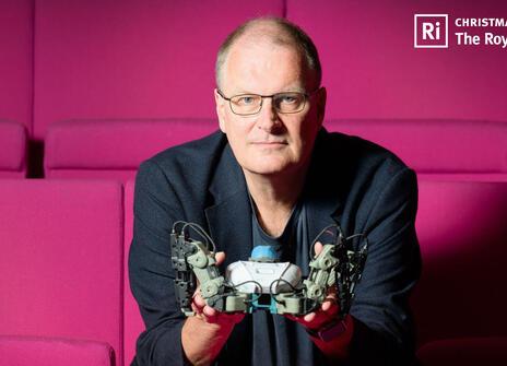 Mike looks into the camera while holding a small robot, sitting on red seats in our Theatre