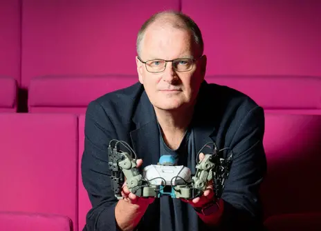Mike looks into the camera while holding a small robot, sitting on red seats in our Theatre