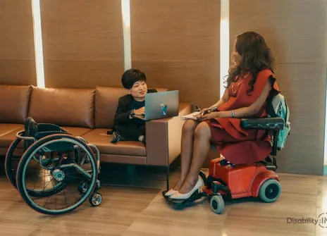 A woman is seated on a sofa, facing another woman sitting in a wheelchair. 