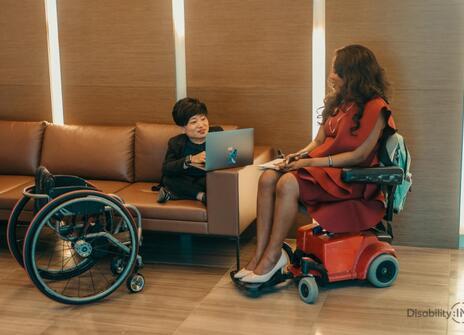 A woman is seated on a sofa, facing another woman sitting in a wheelchair. 