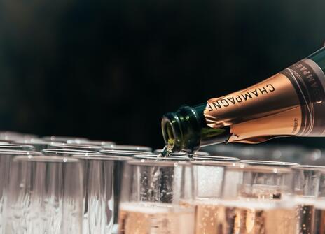 An open bottle of champagne is pouring champagne into lots of flute glasses.